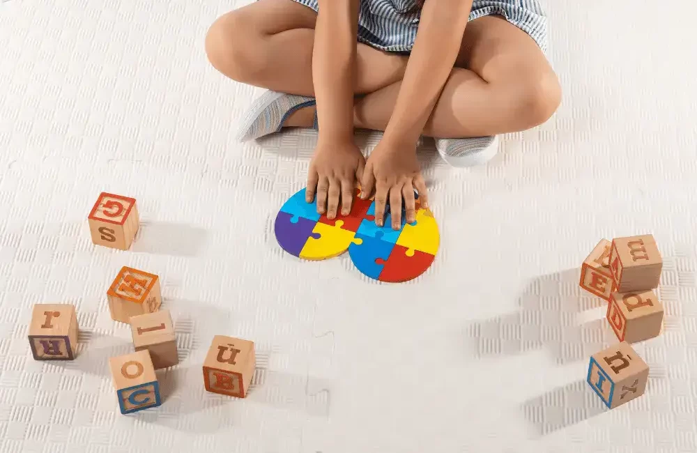 Criança autista sentada no chão com quebra-cabeça colorido em formato de coração montado e dados com letras do alfabeto espalhados no chão ao seu redor.