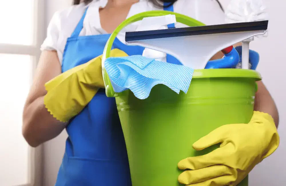 Empregada doméstica utilizando luva nitrílica e carregando balde com produtos de limpeza.